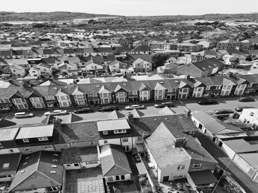 A black and white aerial photograph showing a coastal town with a mixture of residential and commercial buildings. In the foreground, there is a parking lot filled with cars adjacent to a modern flat-